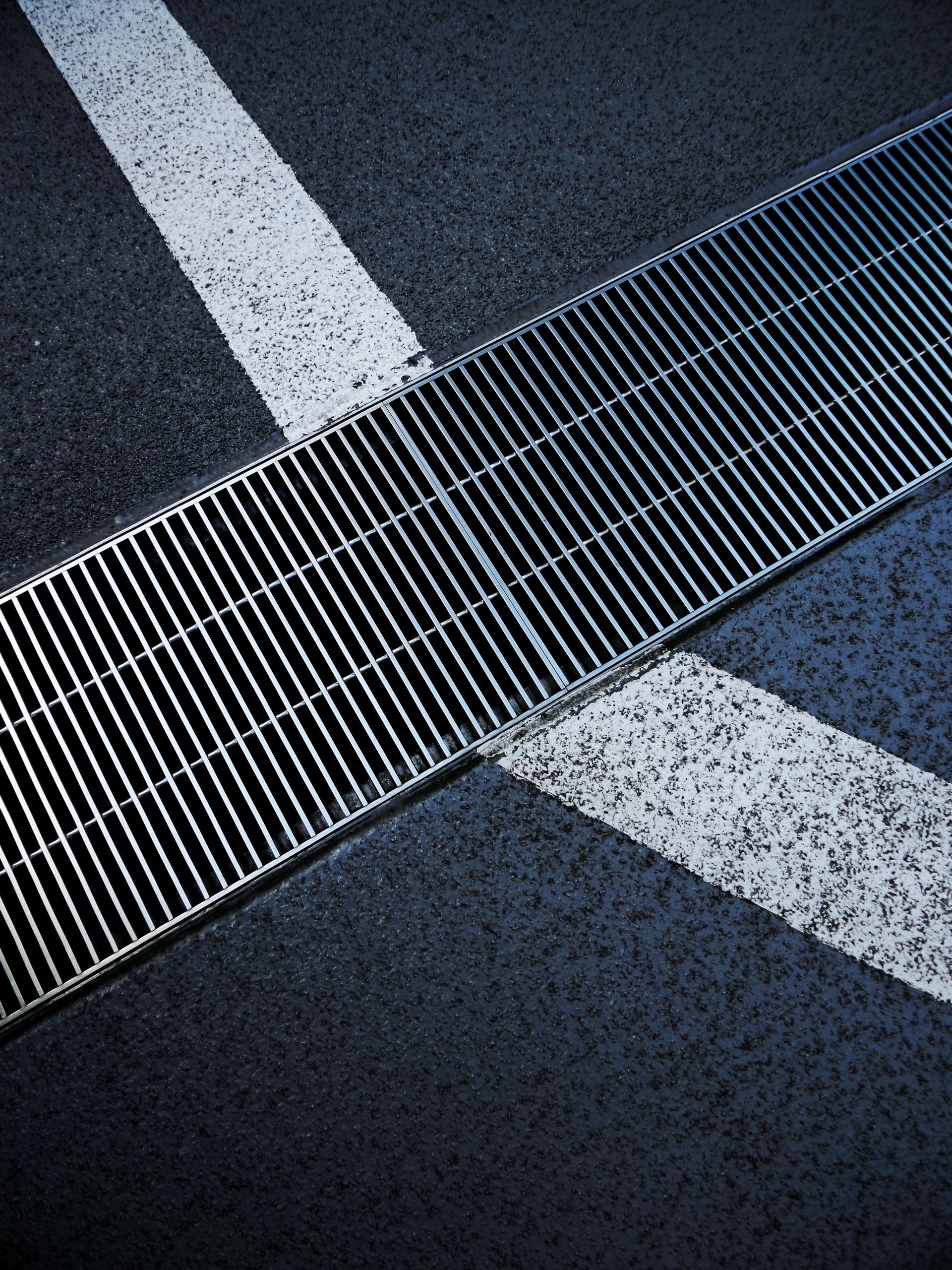 Metallic street grate intersecting white road markings on a textured asphalt surface.
